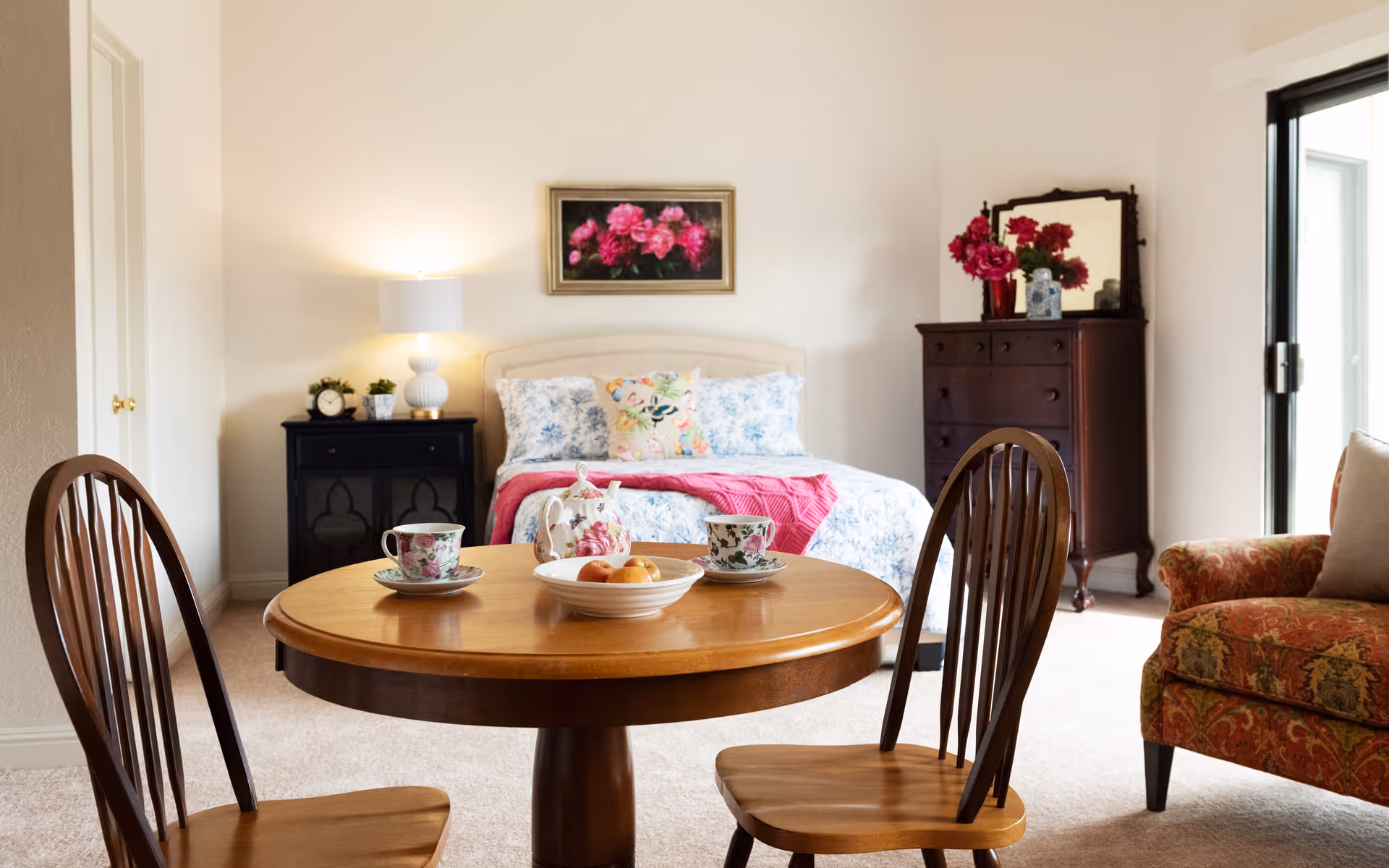 A cozy room featuring a round wooden table set with floral teacups and a bowl of fruit in the foreground. Behind the table is a bed with floral bedding and a pink throw, flanked by a black nightstand with a lamp and clock on the left and a dark wooden dresser with a mirror and red flowers on the right. To the right of the dresser is a patterned armchair and a sliding glass door letting in natural light.