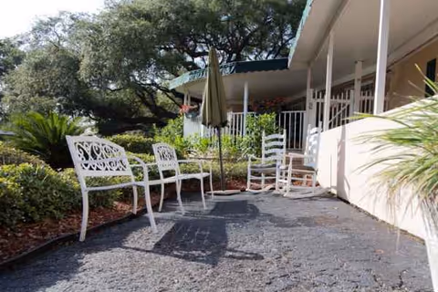 Outdoor patio area with white chairs, table, umbrella, and greenery surrounding a care facility.