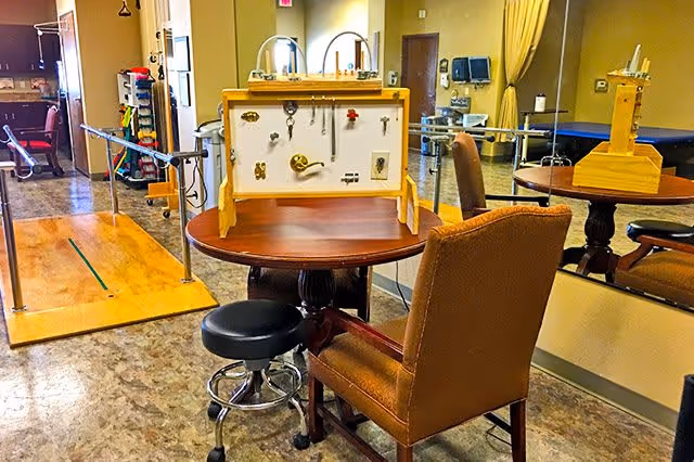 A therapy or rehabilitation room with a wooden table holding a sensory activity board featuring locks, latches, and knobs. There is a brown upholstered chair and a black rolling stool in front of the table. The room has a wooden floor ramp with handrails, exercise equipment, and a large mirror on the wall reflecting part of the room.