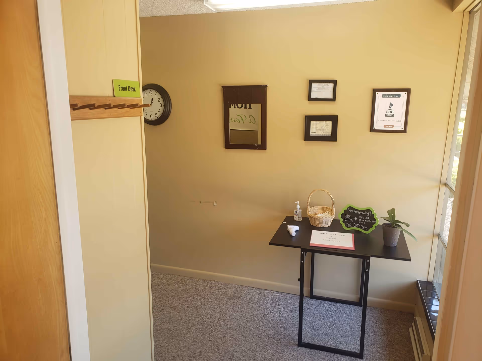 Small reception area with a black table, wall clock, framed certificates, chalkboard sign, and hand sanitizer.