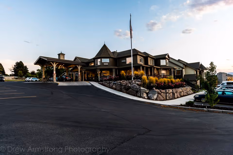 Exterior front of a large assisted living building with a covered entrance, landscaped rock beds, flagpole, and parking lot at dusk.