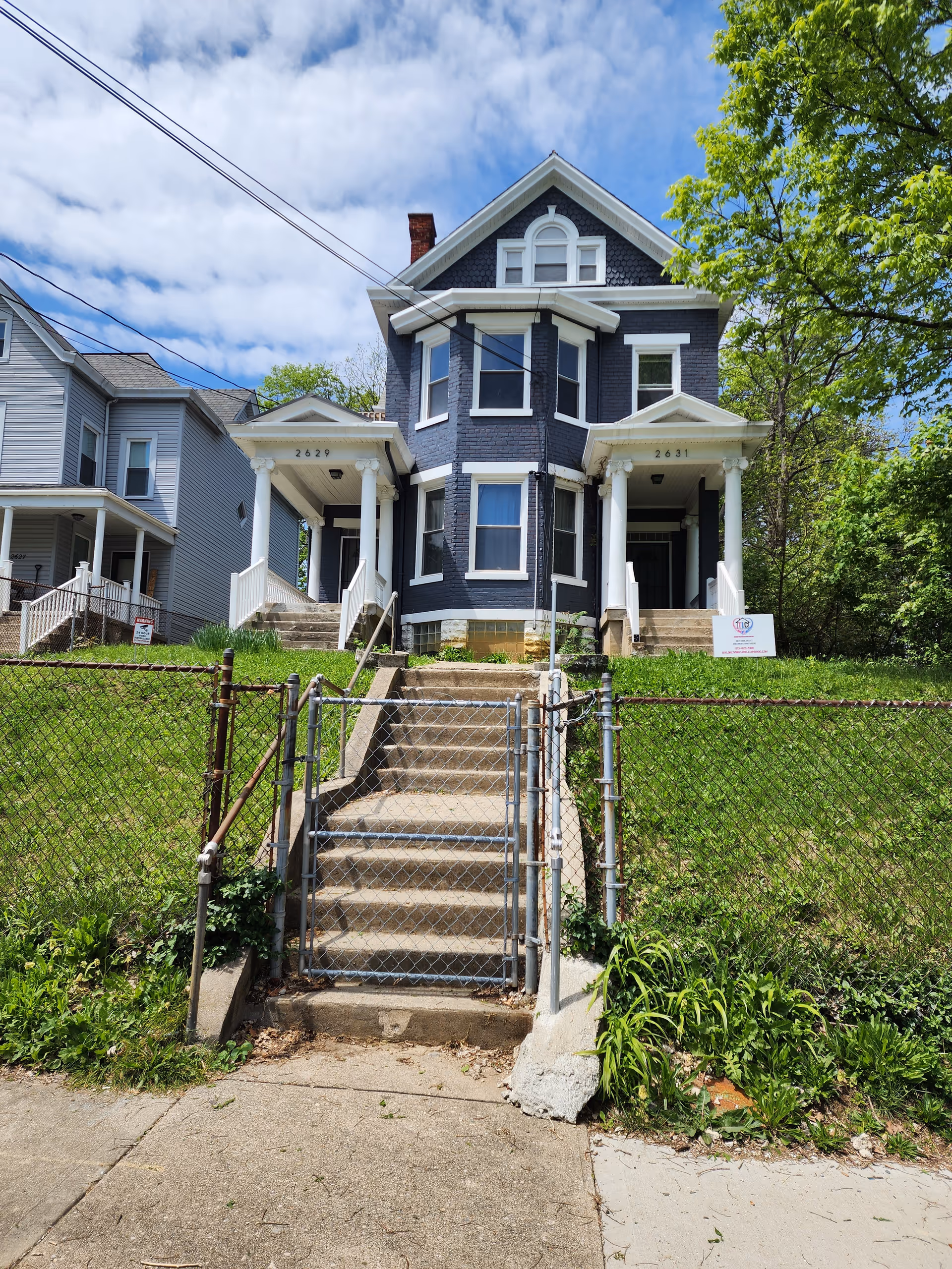 Two-story blue Victorian-style house with front stairs, chain-link fence, and surrounding greenery.