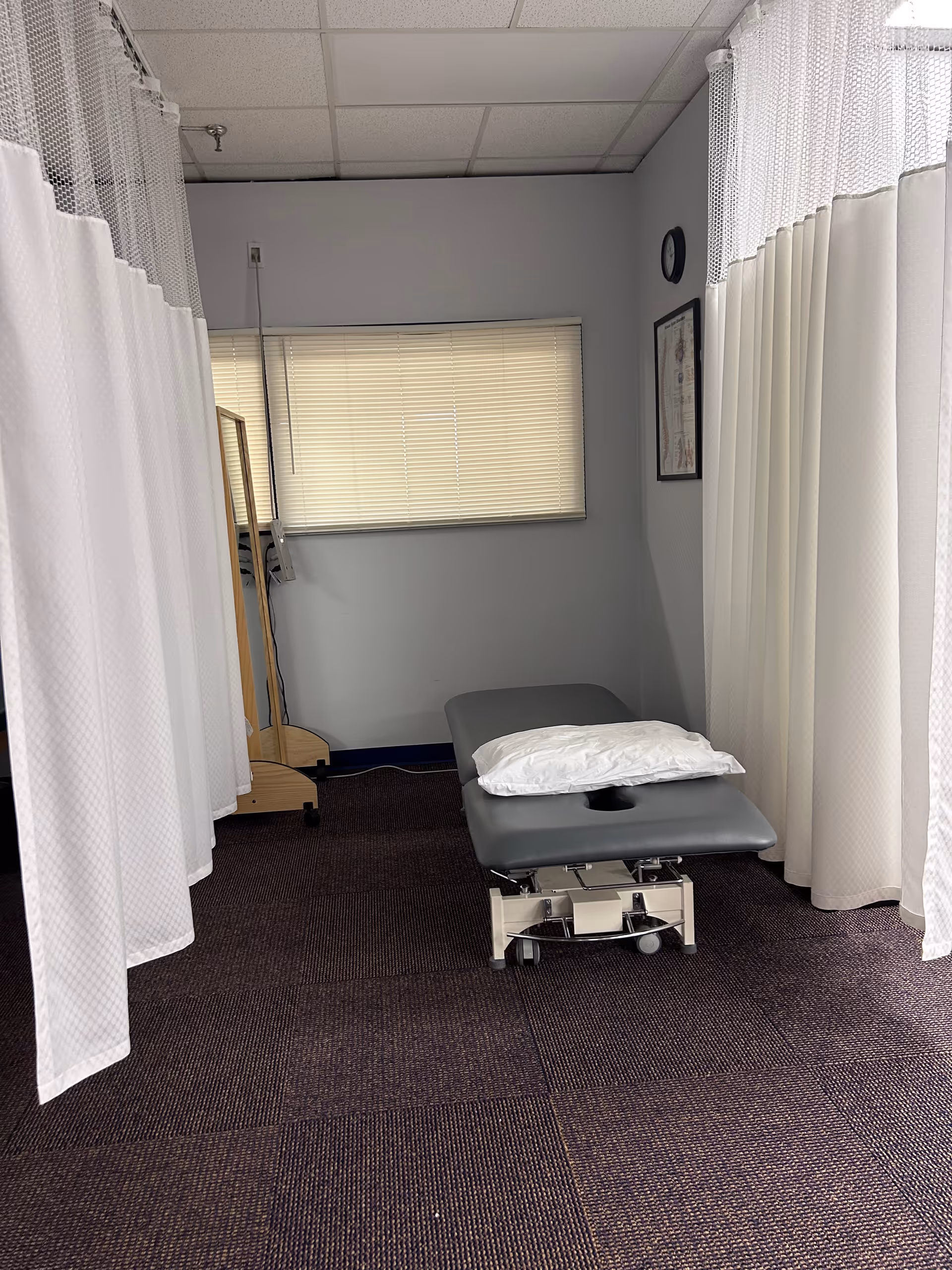 Treatment room with privacy curtains, a padded table, pillow, and window blinds.