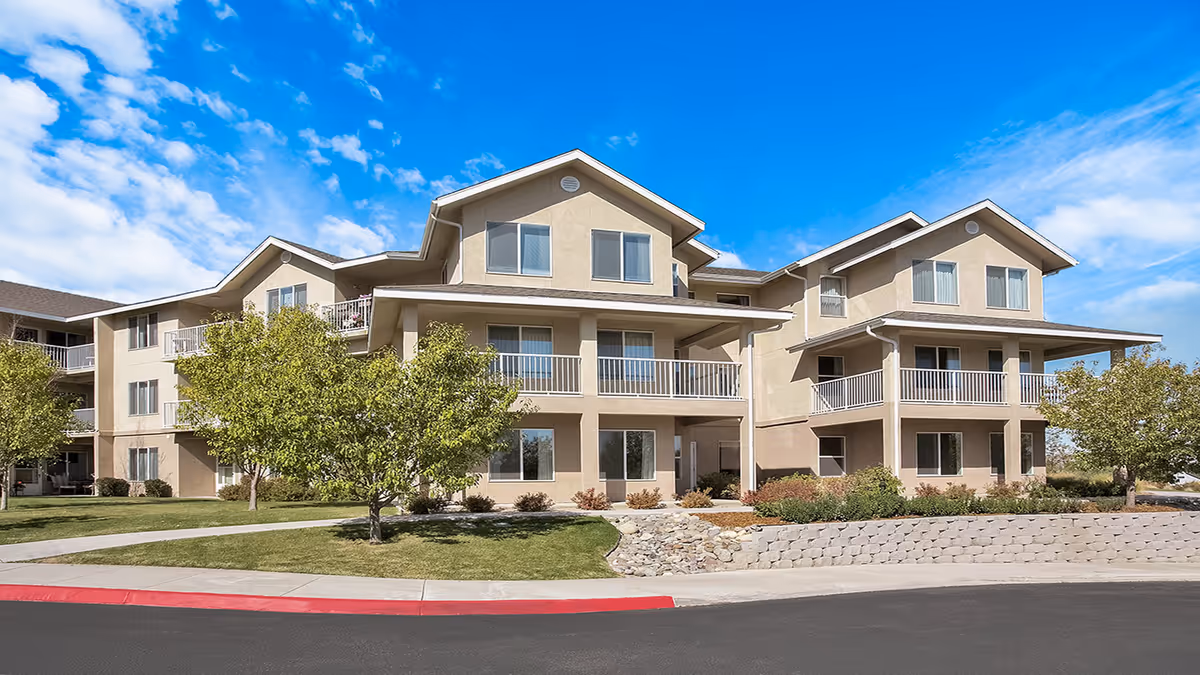 Three-story beige senior living building with balconies, landscaped lawn, trees, and a blue sky.
