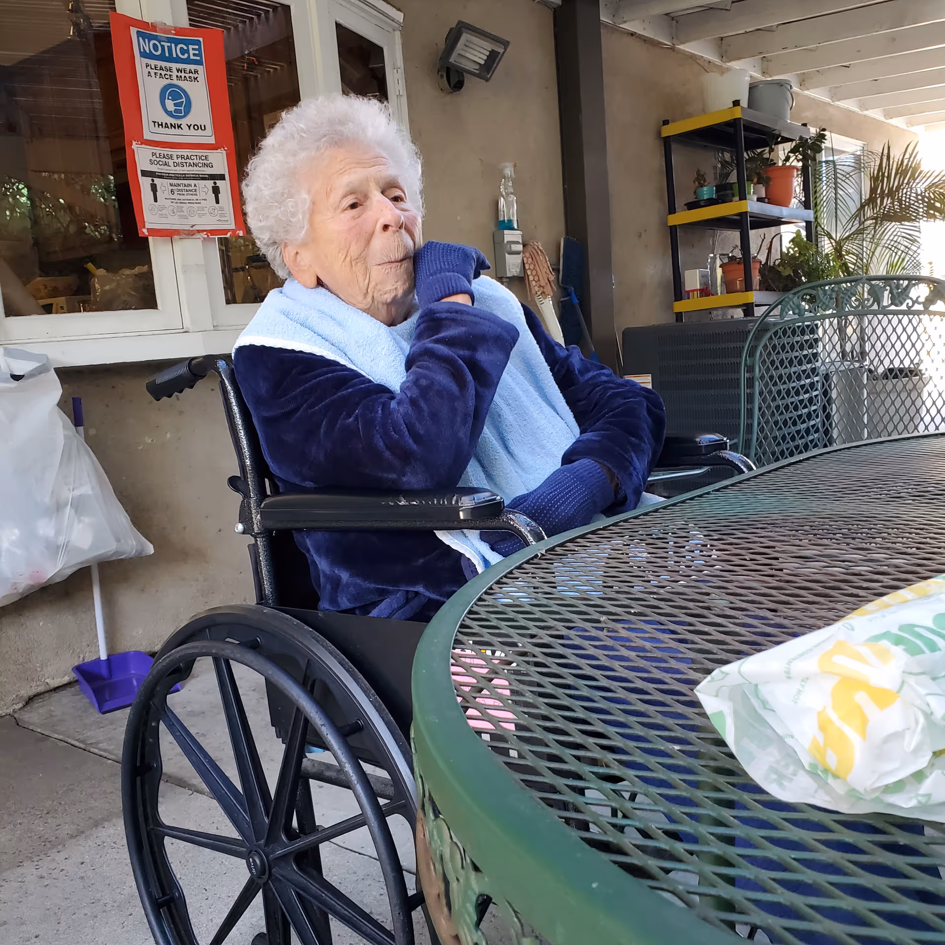 Elderly person in a wheelchair sits outside at a metal table, wearing a blue robe.
