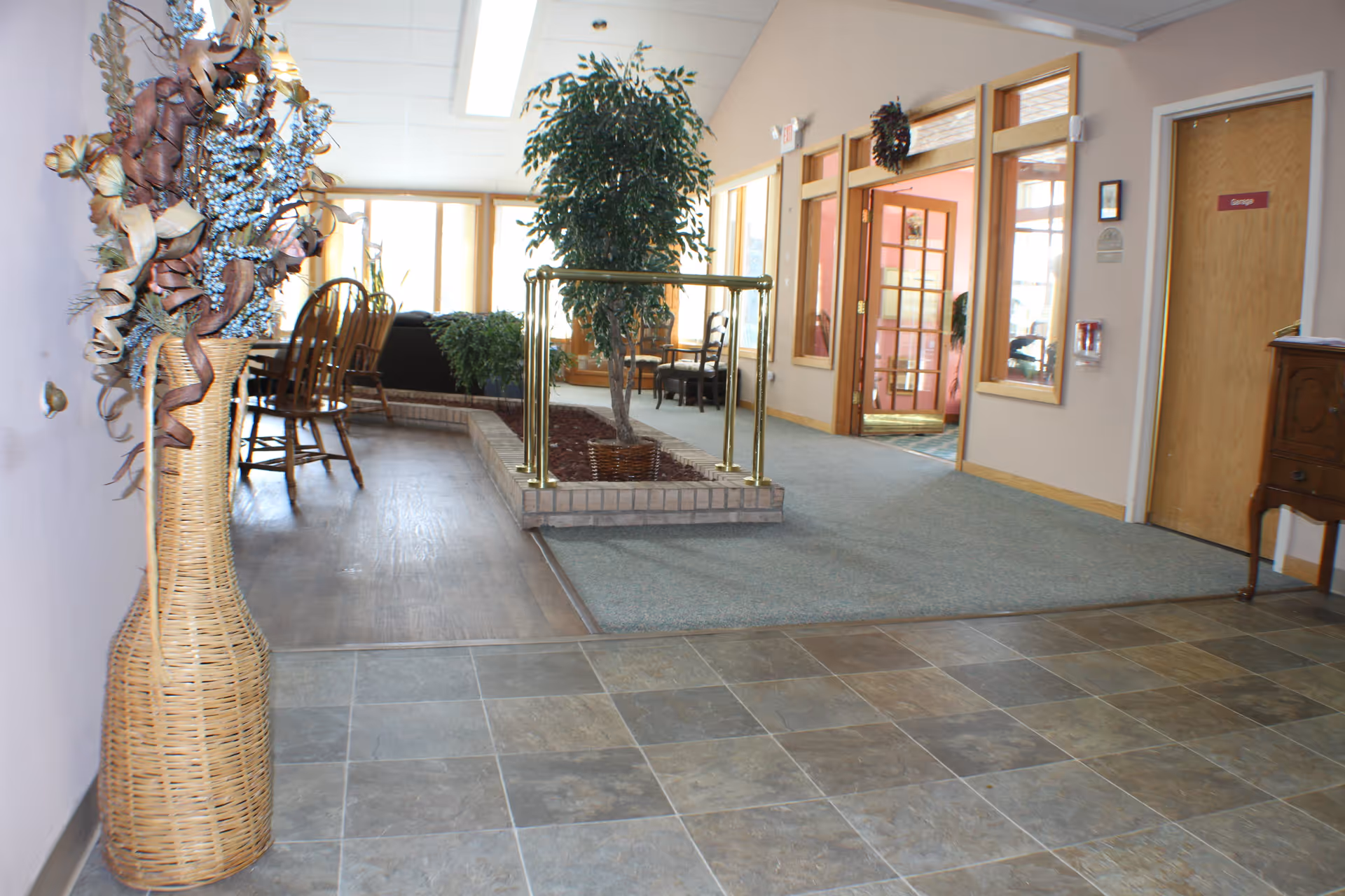 Interior view of a senior living facility hallway with tiled floor in the foreground, a large wicker vase with decorative dried flowers on the left, a small indoor garden with a tree surrounded by a brass railing in the center, and a carpeted area leading to wooden doors and windows on the right. There are chairs and a table visible in the background near large windows.