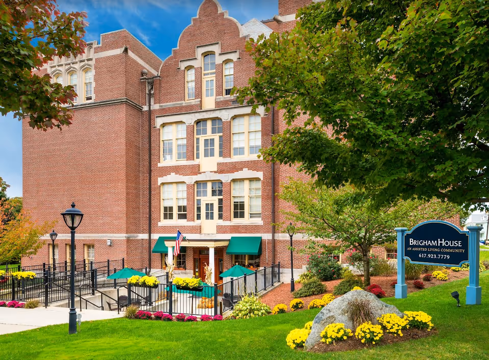 Exterior view of Brigham House, a brick assisted living community building with multiple windows, green awnings, and a landscaped garden featuring yellow and pink flowers, trees, and a large rock. A blue sign with white text displays the facility name and phone number.