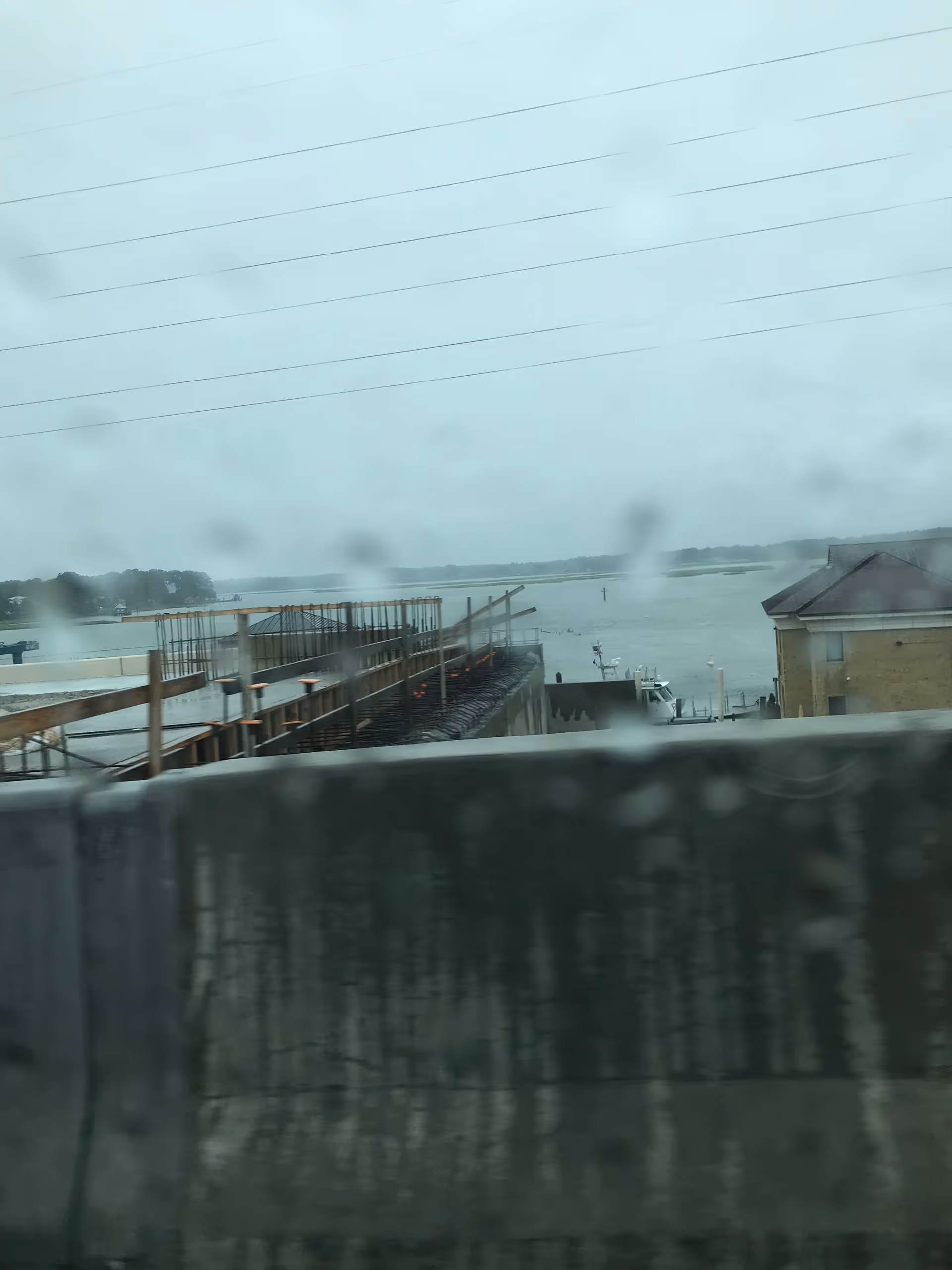 Rainy waterfront scene with construction scaffolding and distant shoreline, photographed through a window.