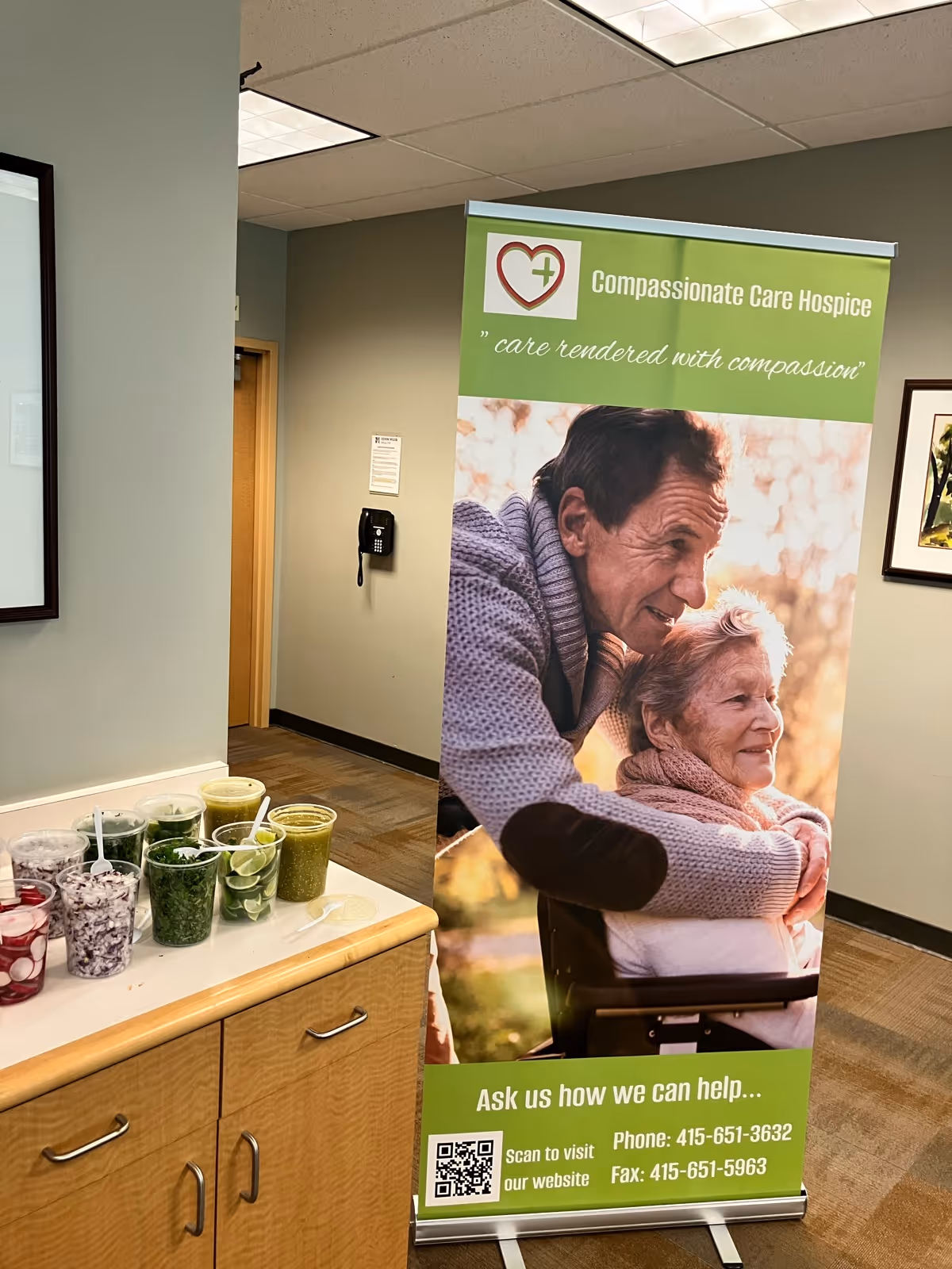 Indoor reception area with a green Compassionate Care Hospice banner beside a drink counter.