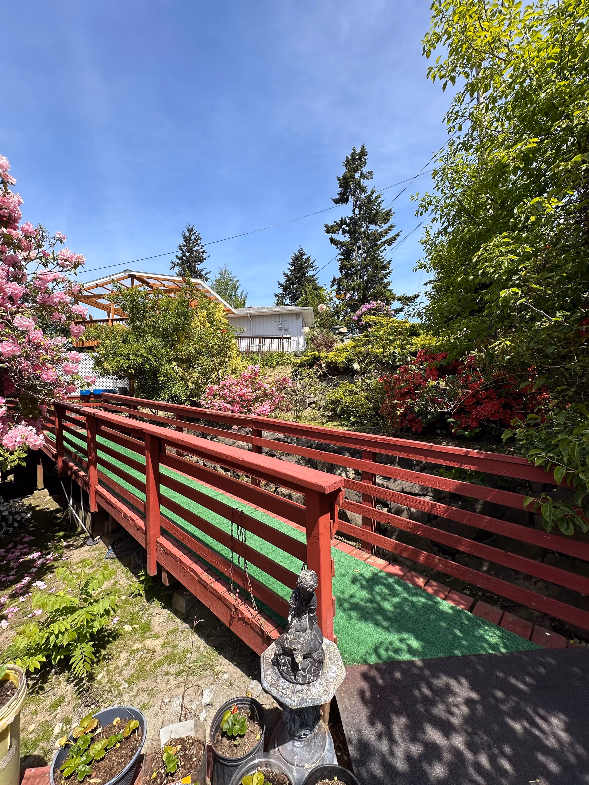 A red wooden footbridge with green carpeting crosses a small garden area filled with various plants and flowers, including pink and red blossoms. In the background, there are tall evergreen trees and a white building with a wooden deck. The sky is clear and blue.