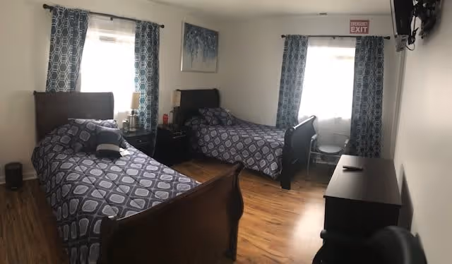 A bedroom with two twin beds featuring dark wooden frames and matching blue patterned bedding. Each bed has a pillow and is positioned against walls with windows covered by blue and white patterned curtains. Between the beds is a small nightstand with a lamp and some items on it. The room has wooden flooring, a dresser with a TV mounted on the wall above it, and an exit sign above one window.