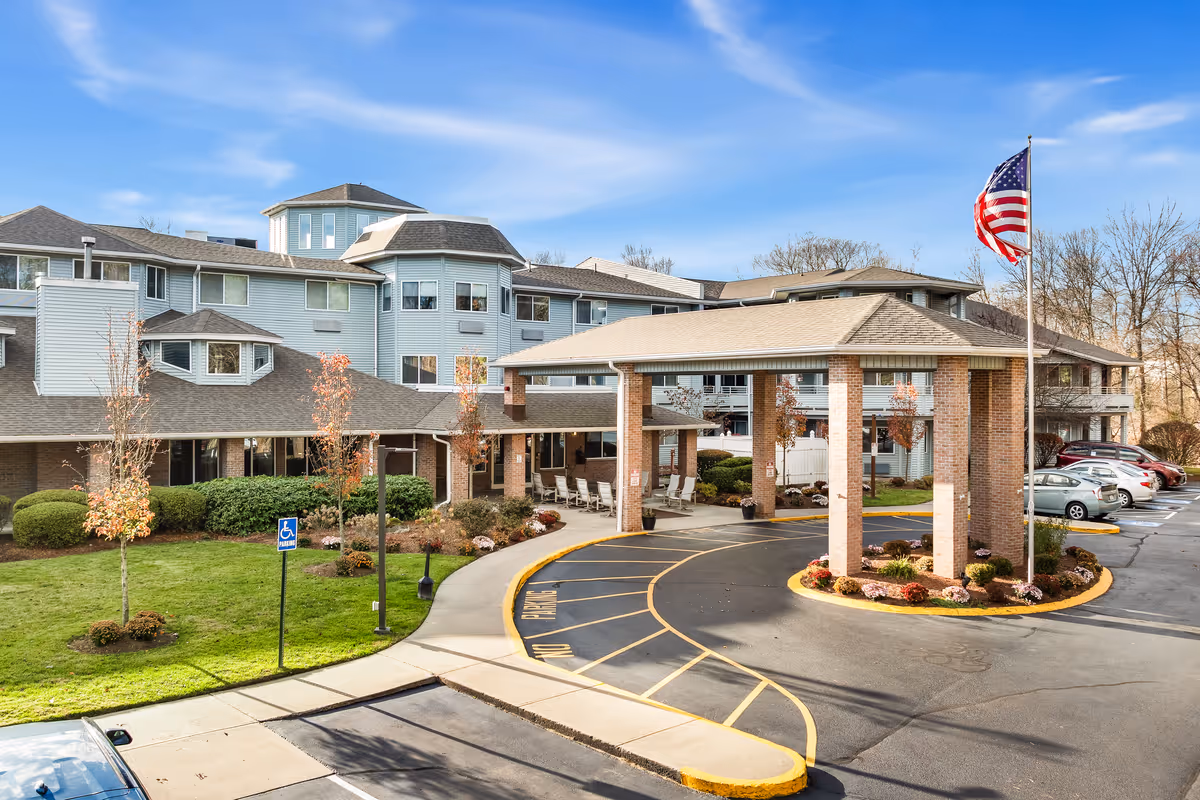 Exterior view of a senior living community building with a covered entrance driveway, landscaped greenery, several parked cars, and an American flag on a flagpole against a blue sky.