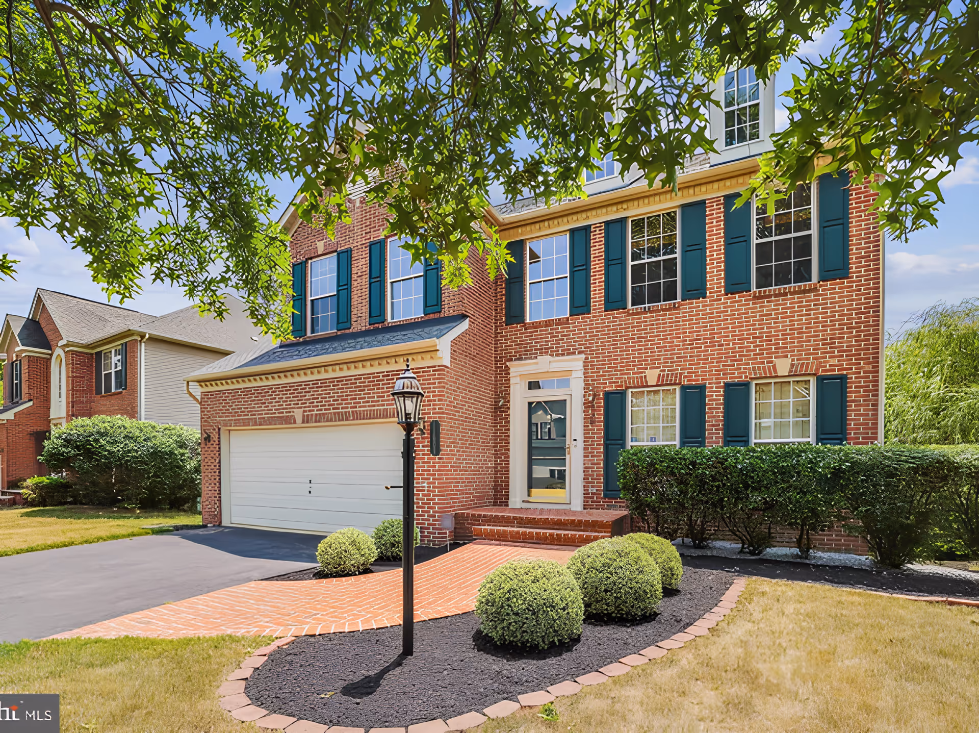 Front exterior view of a two-story brick house with green shutters and a white garage door. The house has a brick walkway leading to the front door, neatly trimmed bushes, a black lamp post, and a well-maintained lawn with trees providing shade.