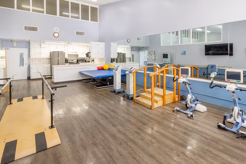 A rehabilitation room with exercise equipment including stationary bikes, a wooden step platform with handrails, parallel bars for walking practice, and a padded therapy table. The room has a large mirror on one wall, white cabinets, and a kitchen area in the background.