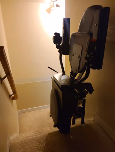 An indoor stairlift chair is mounted on a railing in a narrow carpeted stairway.
