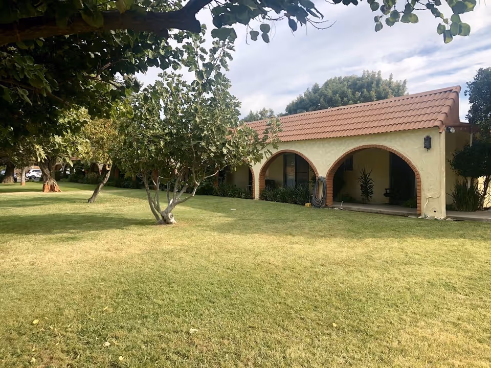 Single-story stucco building with arched walkways, a red tile roof and a grassy front lawn with trees.