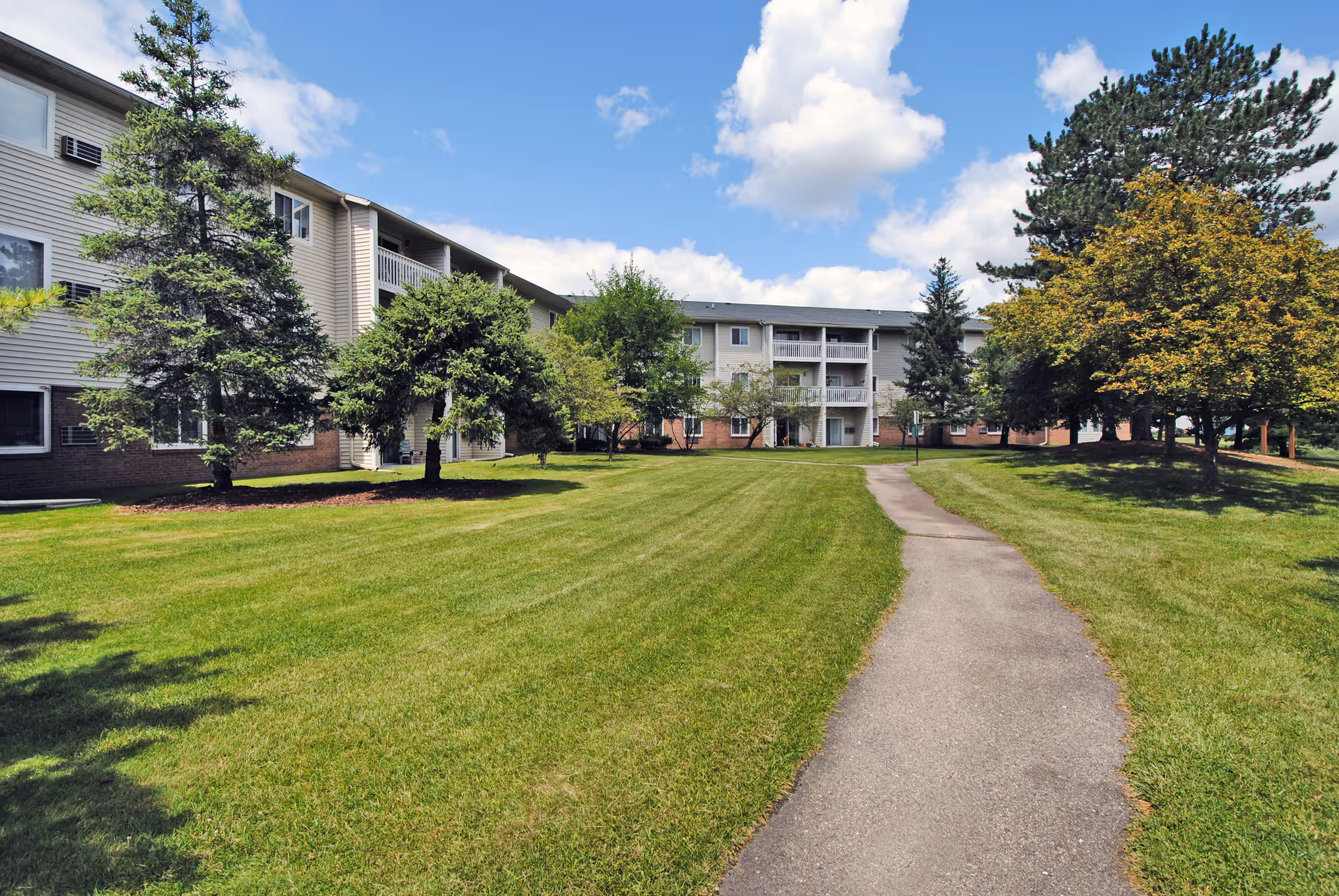 Paved walkway through a grassy campus toward a multi-story senior living building with trees and balconies