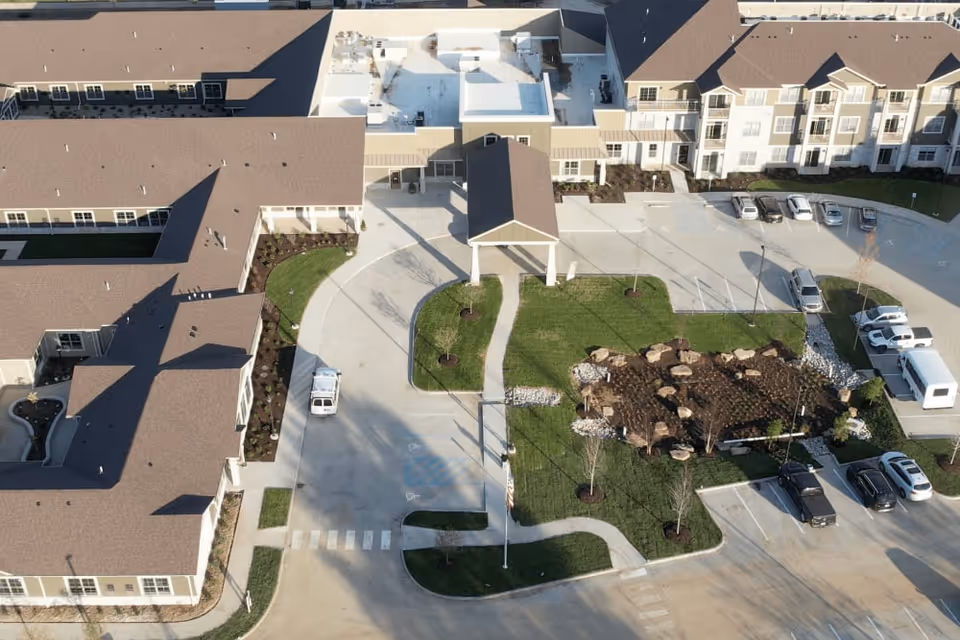 Aerial view of The Westbury Senior Living facility showing multiple connected buildings with brown roofs, a central driveway, landscaped green areas with small trees and rocks, and several parked vehicles.