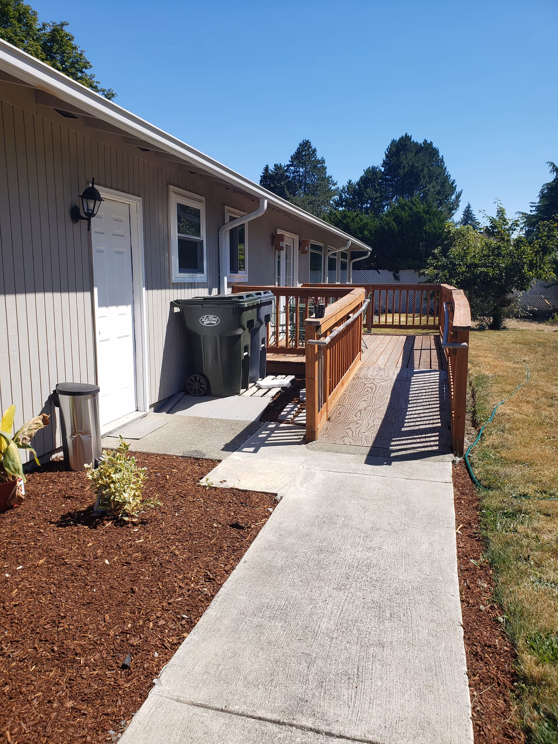 Exterior view of a single-story senior home with a wooden ramp and trash bins.