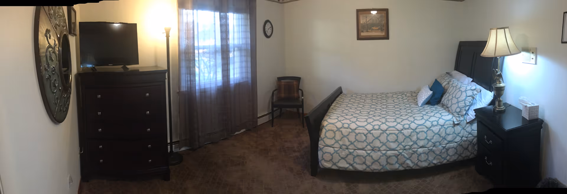 Senior living bedroom featuring a bed, dresser, lamps, TV, and window with curtains.