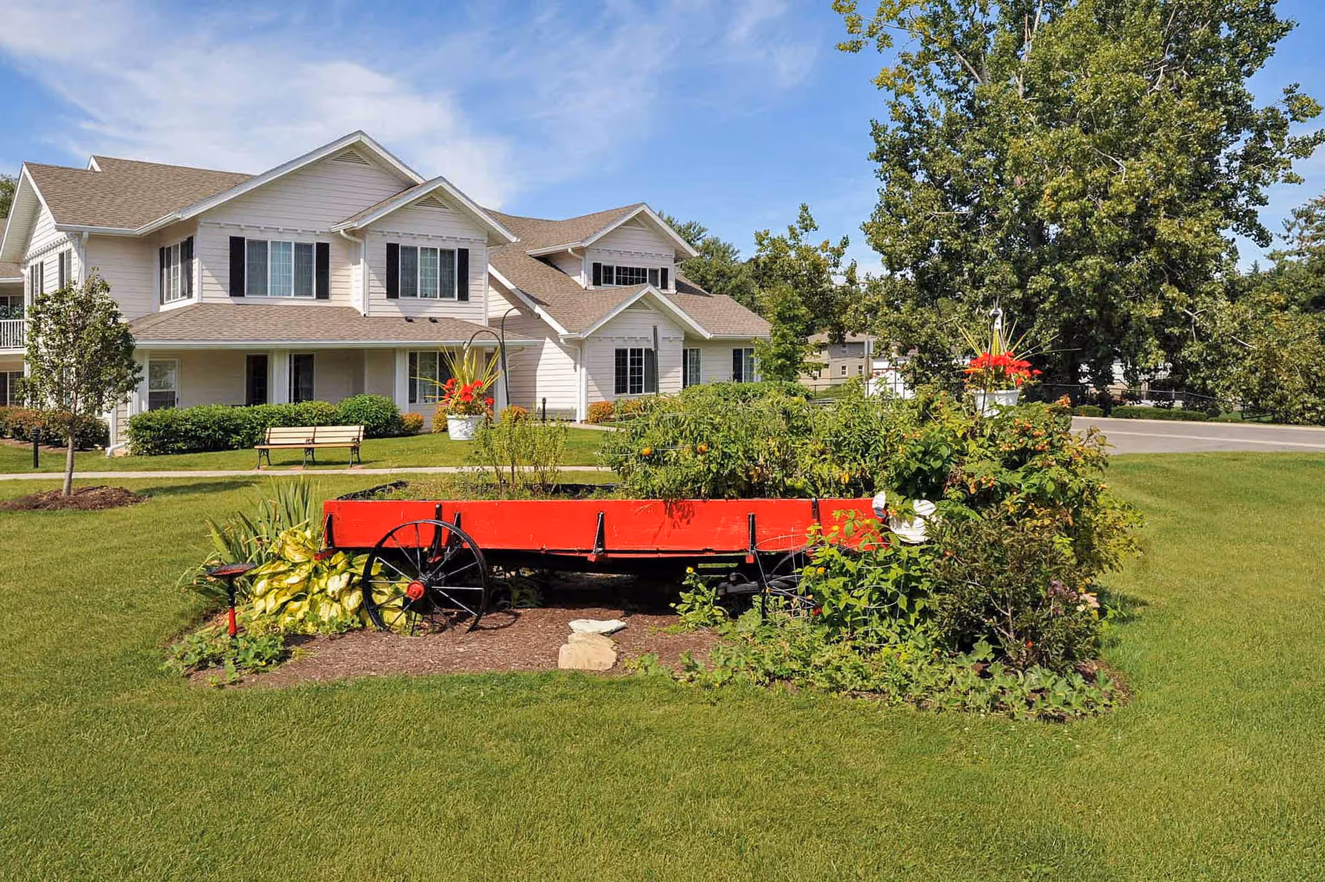 White two-story residential building with a manicured lawn and a red wagon planter filled with plants in the foreground.