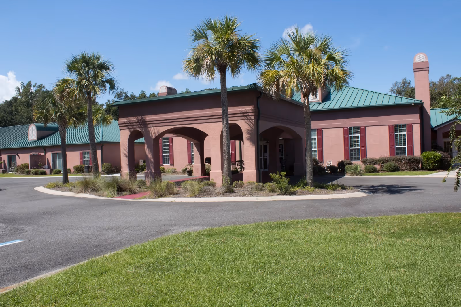 Exterior view of Magnolia Manor of St. Marys, showing a single-story building with a green metal roof and pinkish walls. The entrance features a covered portico supported by arches, surrounded by palm trees and landscaped greenery under a clear blue sky.
