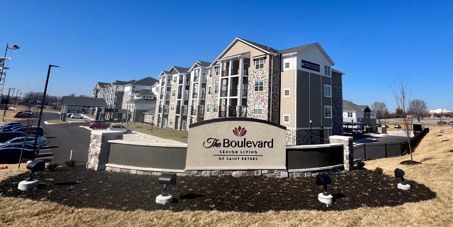 Front exterior of The Boulevard Senior Living of Saint Peters with its entrance sign, multi-story building, parking lot and grounds under a clear blue sky.