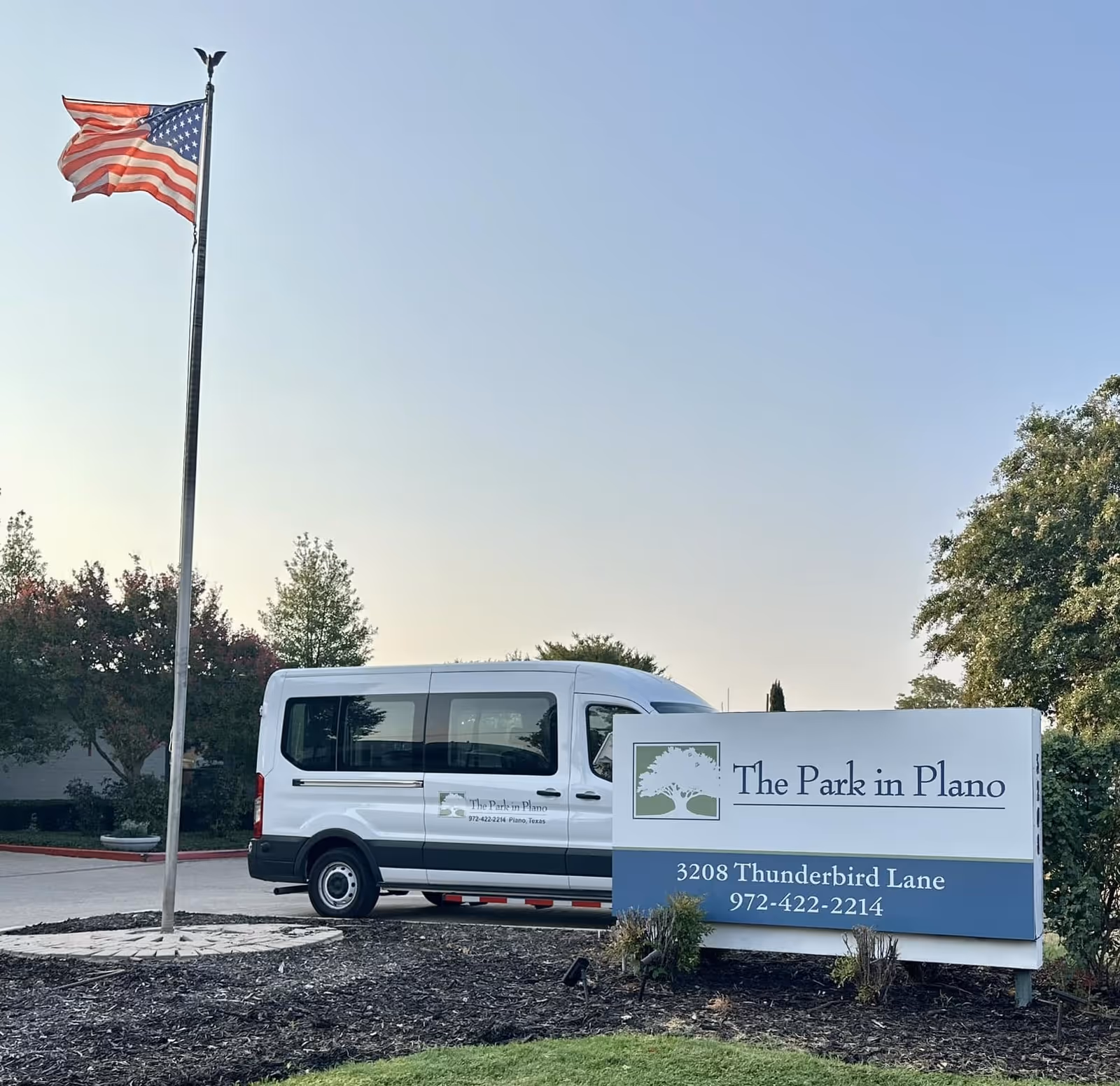 An outdoor scene showing a white shuttle van parked behind a sign for The Park in Plano, located at 3208 Thunderbird Lane with phone number 972-422-2214. An American flag on a flagpole is flying to the left, and trees and bushes surround the area under a clear sky.