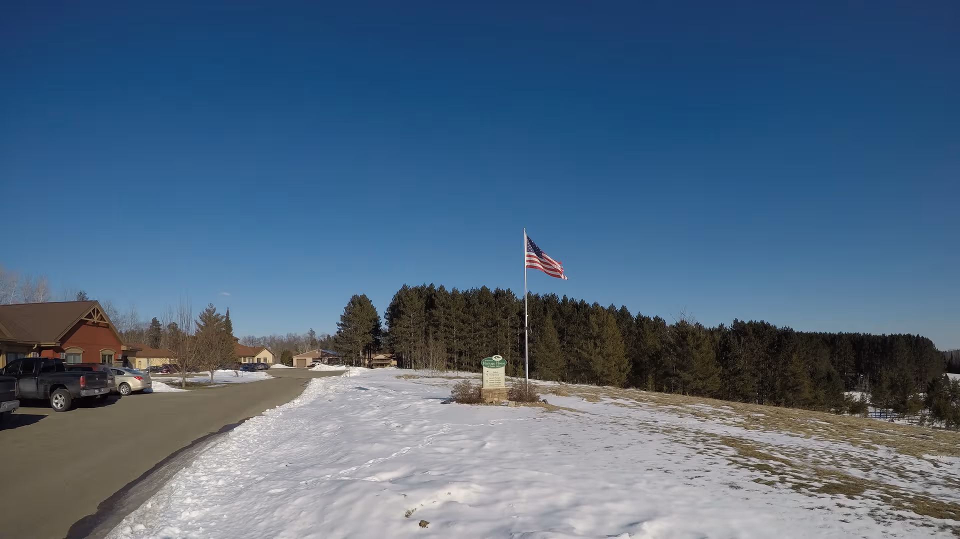 Snow on ground with parked cars, a flagpole, and a building front under a blue sky.