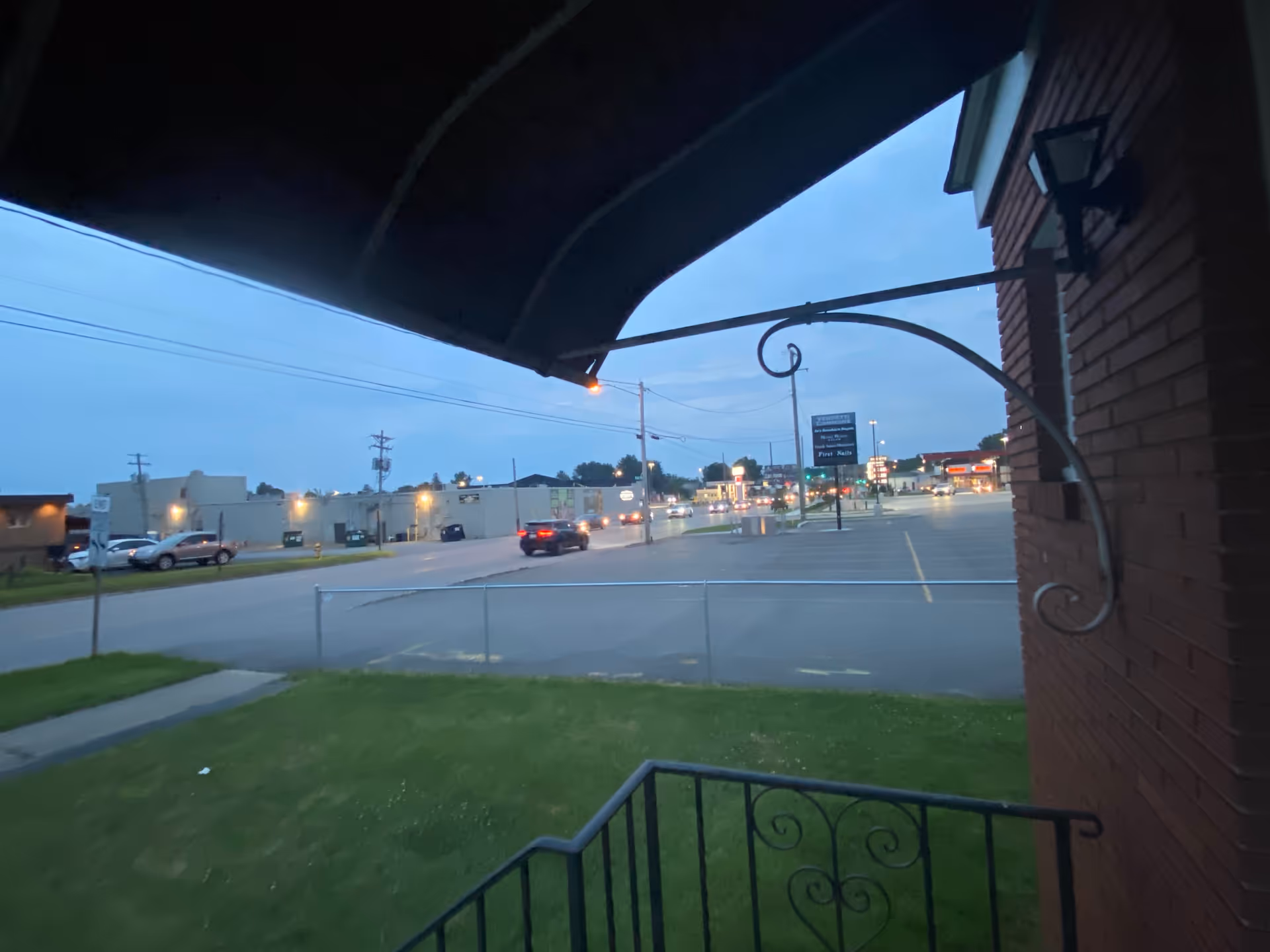 View from a covered porch shows a brick building, lawn, and a distant parking lot.