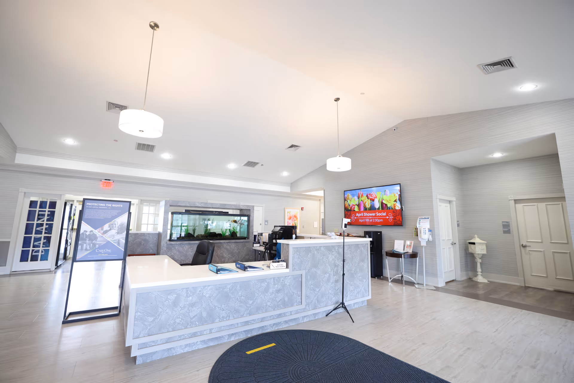 Bright senior living lobby with a front desk, aquarium, TV, and seating area.