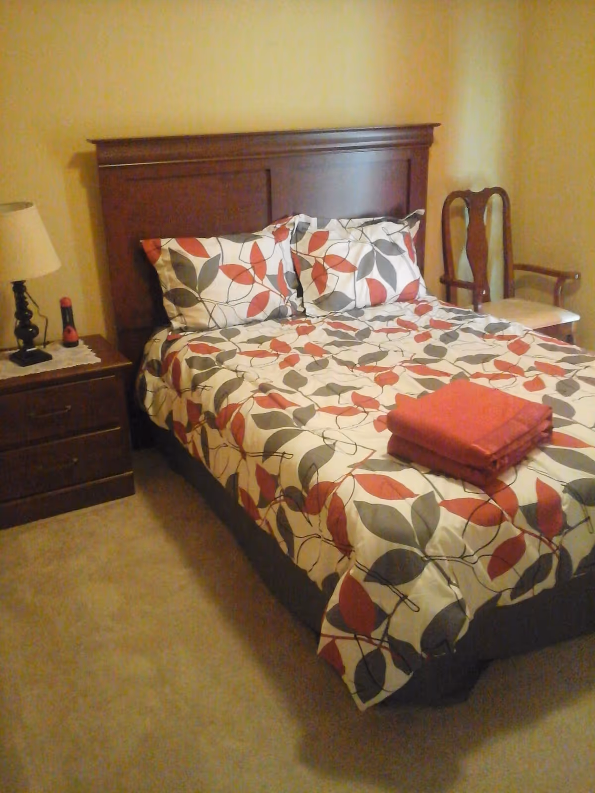 A bedroom with a bed that has a wooden headboard and floral patterned bedding in red, gray, and white colors. There are two pillows matching the bedding and a folded red blanket on the bed. To the left of the bed is a wooden nightstand with a lamp and a small red flashlight. To the right of the bed is a wooden chair with a cushioned seat. The walls are painted light yellow and the floor is carpeted.
