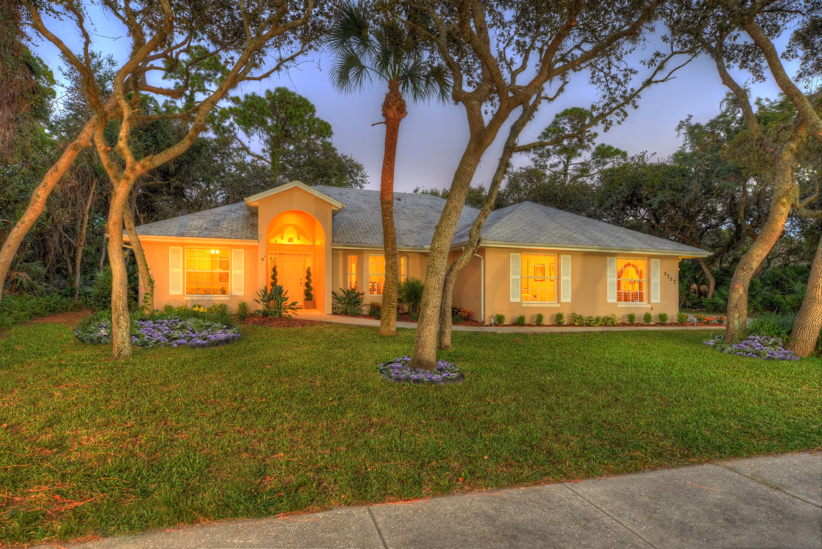 Single-story house with illuminated windows, a manicured lawn and trees in front at dusk.