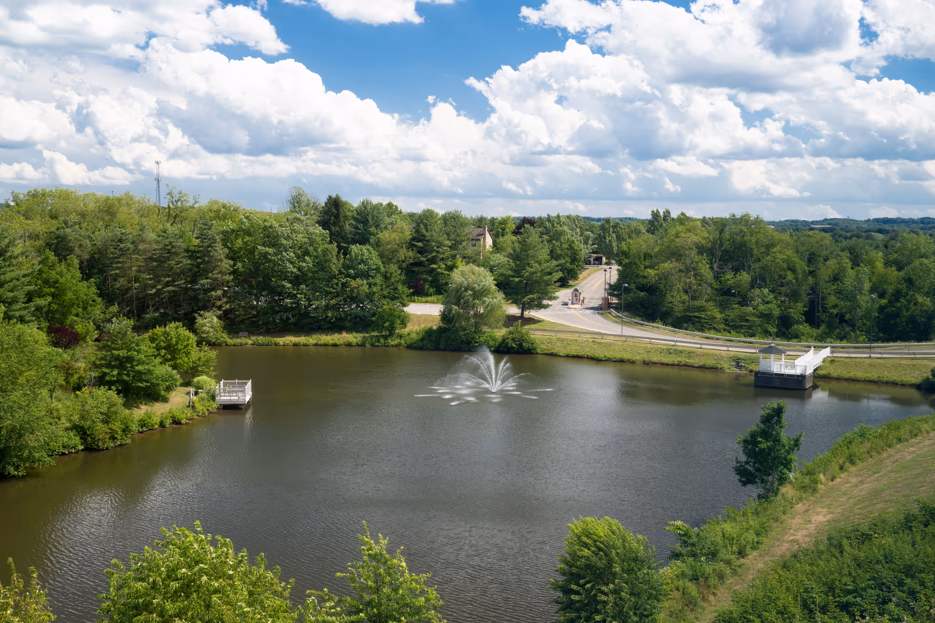 A serene lake surrounded by trees with a central fountain and a distant road.