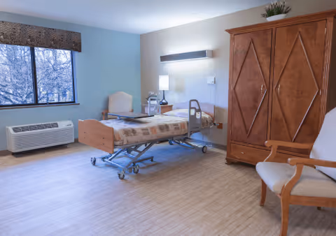 A bright patient bedroom with an adjustable hospital bed, bedside table and lamp, wooden armoire, and seating by a window.