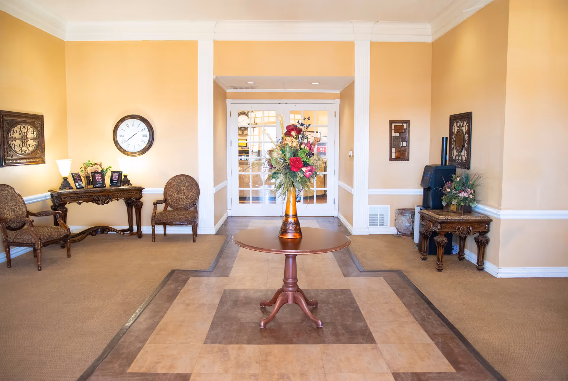 Bright reception lobby with a round center table topped by a floral arrangement, seating and decorative side tables against warm beige walls and glass double doors.