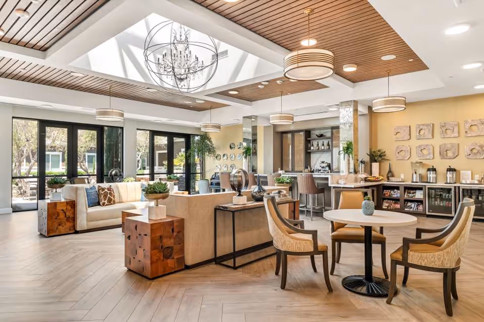 A bright and spacious senior living facility common area with modern decor, featuring a seating area with sofas and wooden side tables, a round table with four chairs, large windows letting in natural light, a chandelier hanging from a skylight, and a refreshment area with coffee dispensers and snacks.