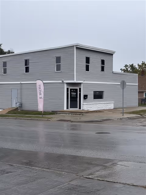 Gray two-story building exterior with front entrance, flag, and street in front.