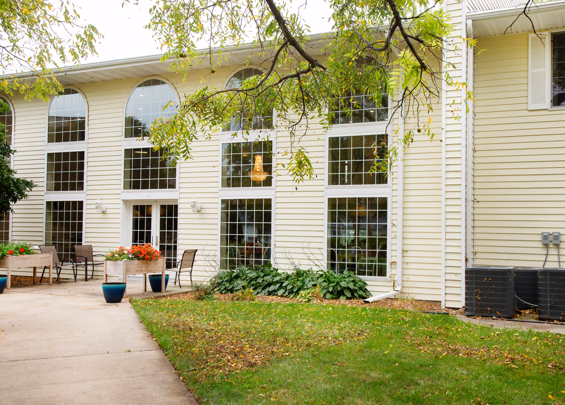 Exterior view of Charleston House Assisted Living building with large arched windows, a concrete walkway, outdoor seating with chairs and flower planters, green grass, and tree branches overhead.