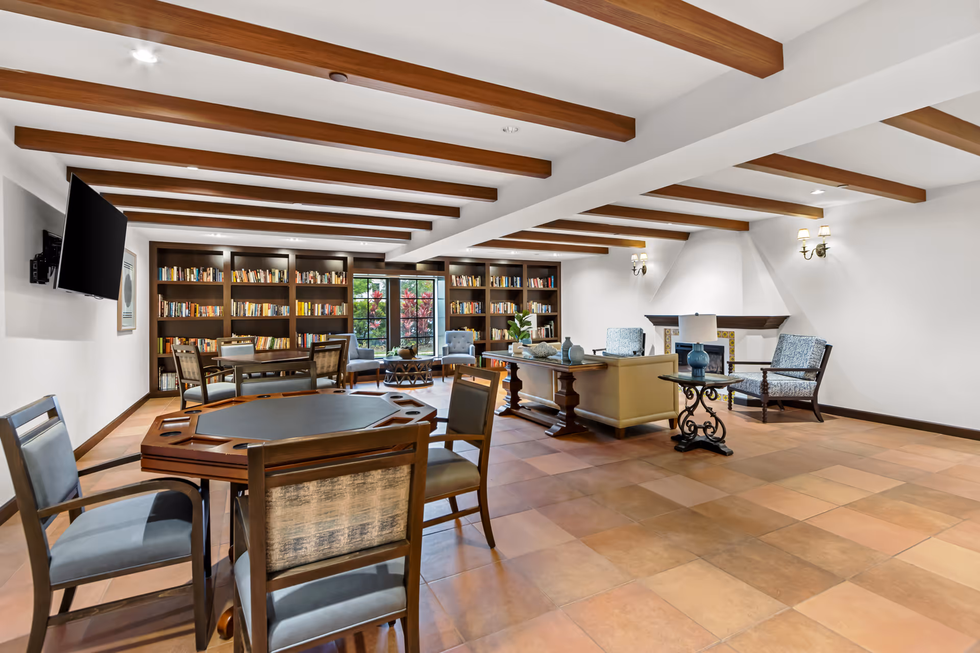 A spacious, well-lit common area with wooden ceiling beams, a tiled floor, and a large bookshelf filled with books along the back wall. The room features a poker table with chairs in the foreground, a seating area with armchairs and a sofa near a fireplace, and a wall-mounted flat-screen TV on the left side. Large windows provide natural light and a view of greenery outside.