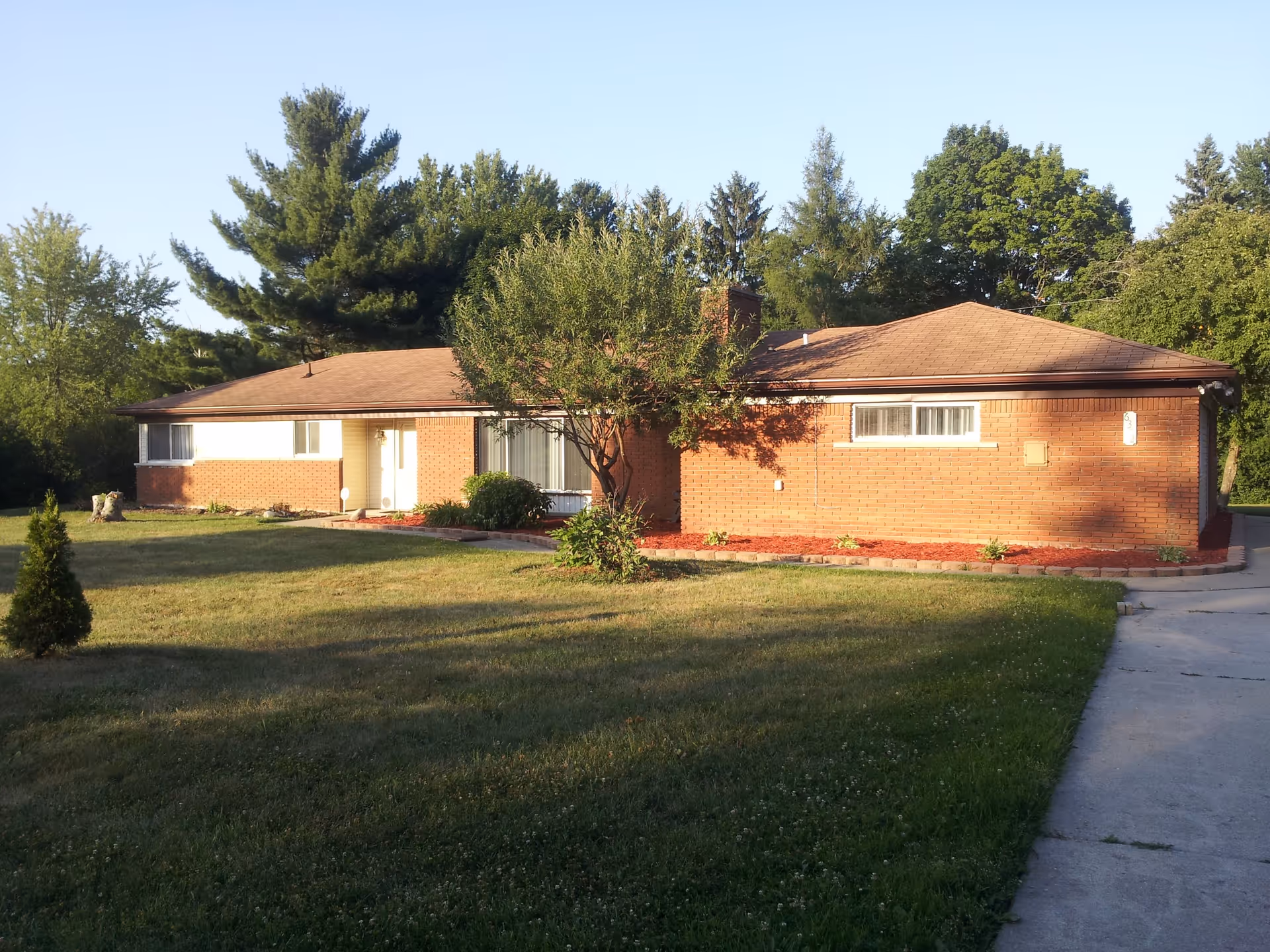 Single-story brick ranch-style house with a front lawn, tree, and driveway in sunlight.