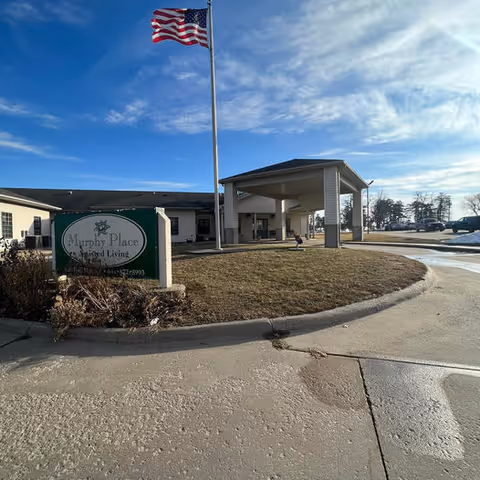 Exterior view of Murphy Place assisted living facility with a covered entrance, an American flag on a flagpole, and a sign displaying the facility's name. The sky is clear with some clouds, and there is a driveway and parking area visible.