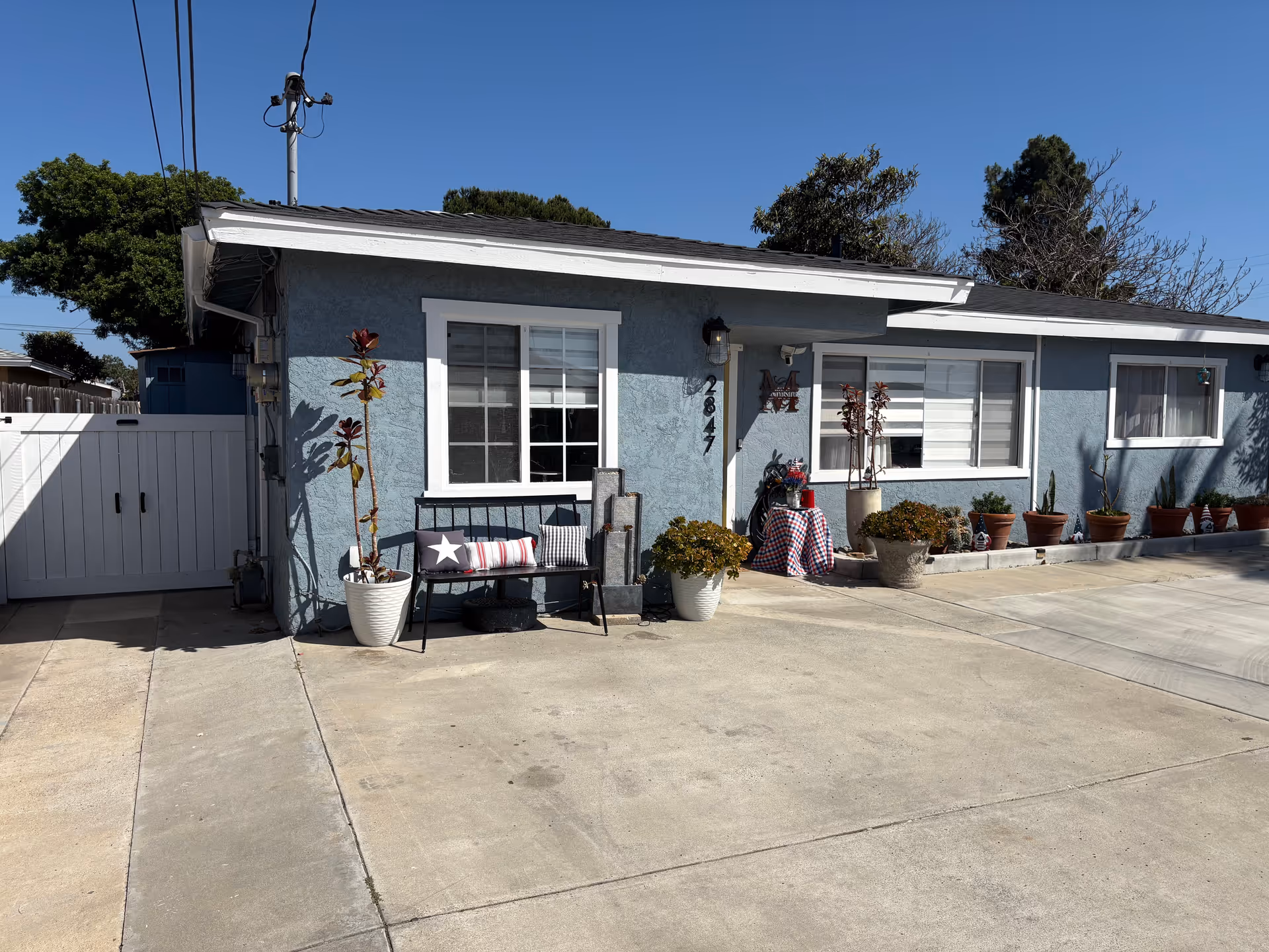 Blue single-story building exterior with multiple potted plants along a concrete driveway and seating.