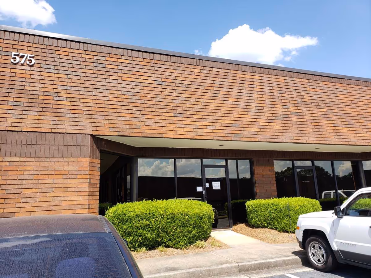 Brick building exterior with glass entrance, trimmed hedges, and parked cars.