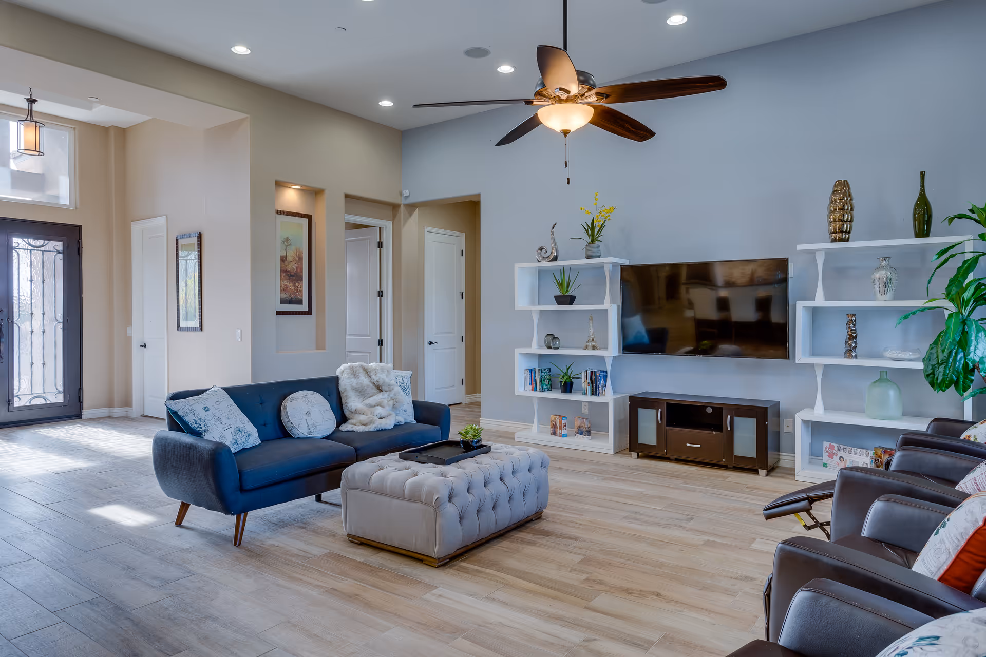 Bright living room with blue sofa, tufted ottoman, TV, white shelves, and ceiling fan