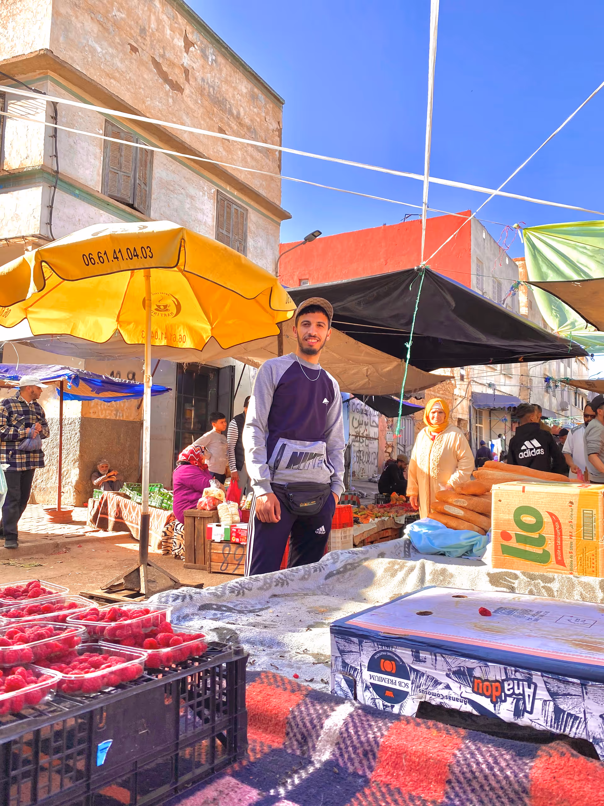 A man in casual clothes stands at an outdoor market with stalls and bright awnings.