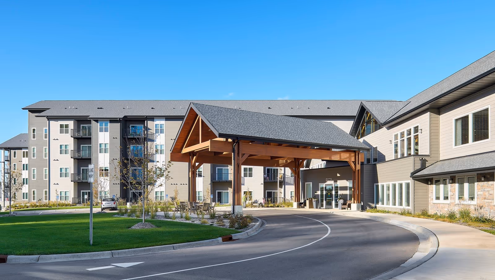 Exterior view of Benedictine Living Community- Shakopee Windermere Way showing a large building with multiple floors, balconies, and a covered entrance with wooden beams. The driveway curves around a landscaped area with grass and small trees under a clear blue sky.