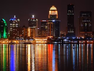 Night skyline of tall buildings along a river with colorful reflections.