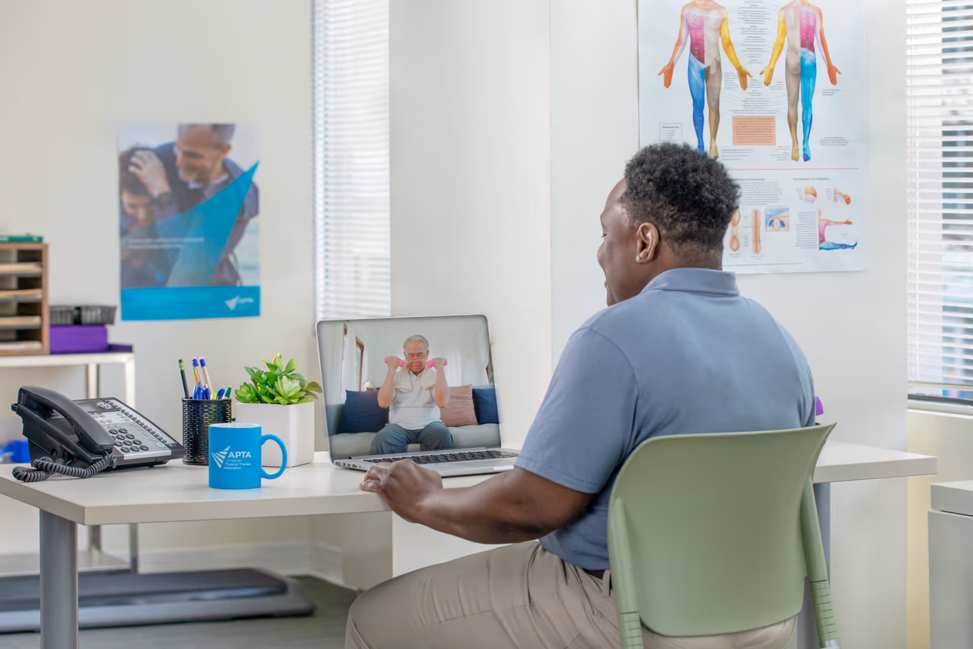 A caregiver sits at a desk in a bright clinic office, video calling a resident.