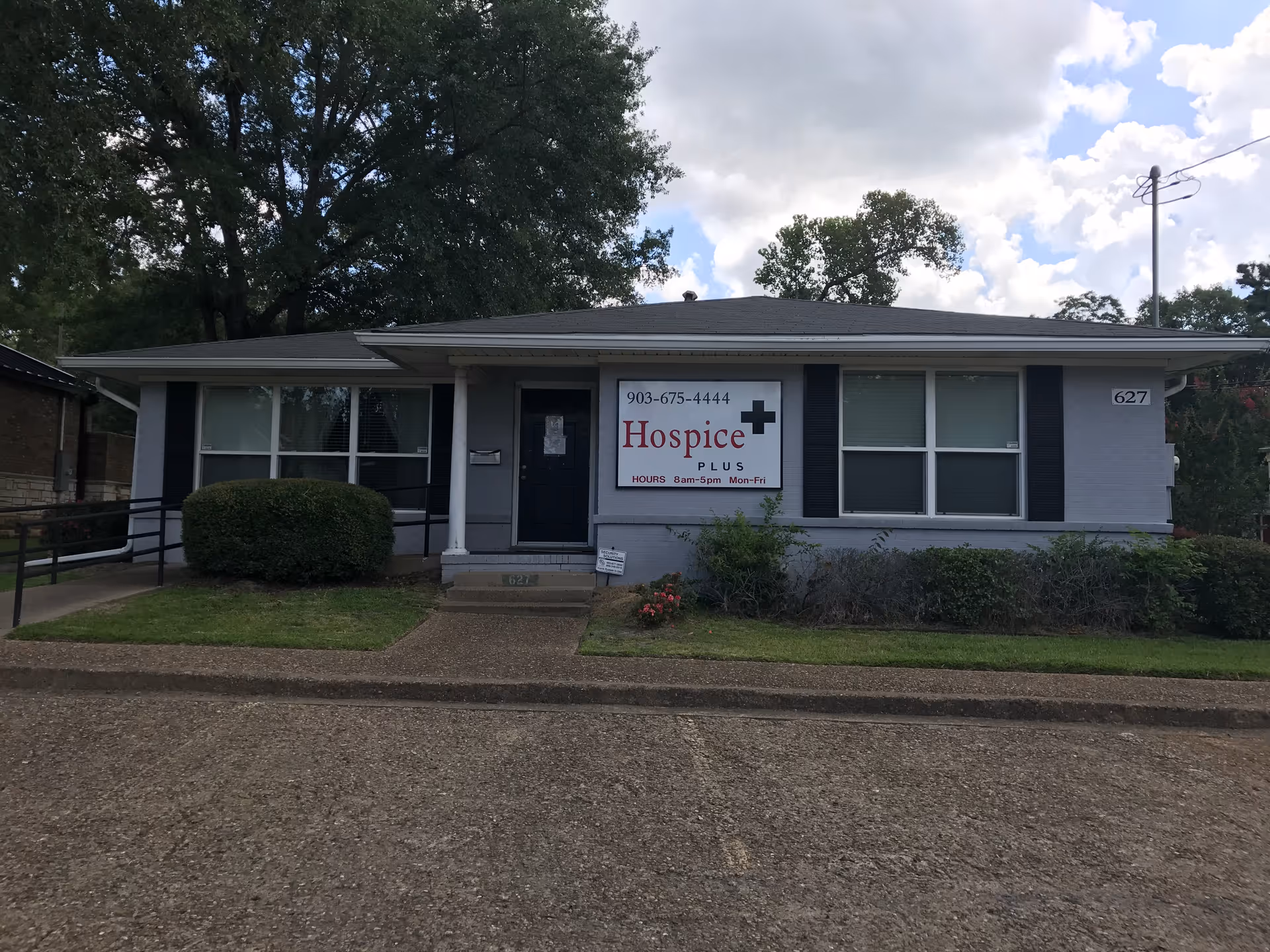 Single-story building with Hospice Plus sign and front entrance, surrounded by shrubs and trees.